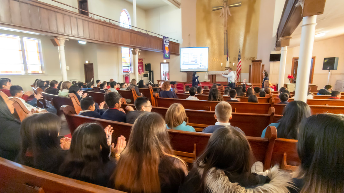 Students sitting and clapping in a chapel service while Principal Carlo Giannotta leads the service